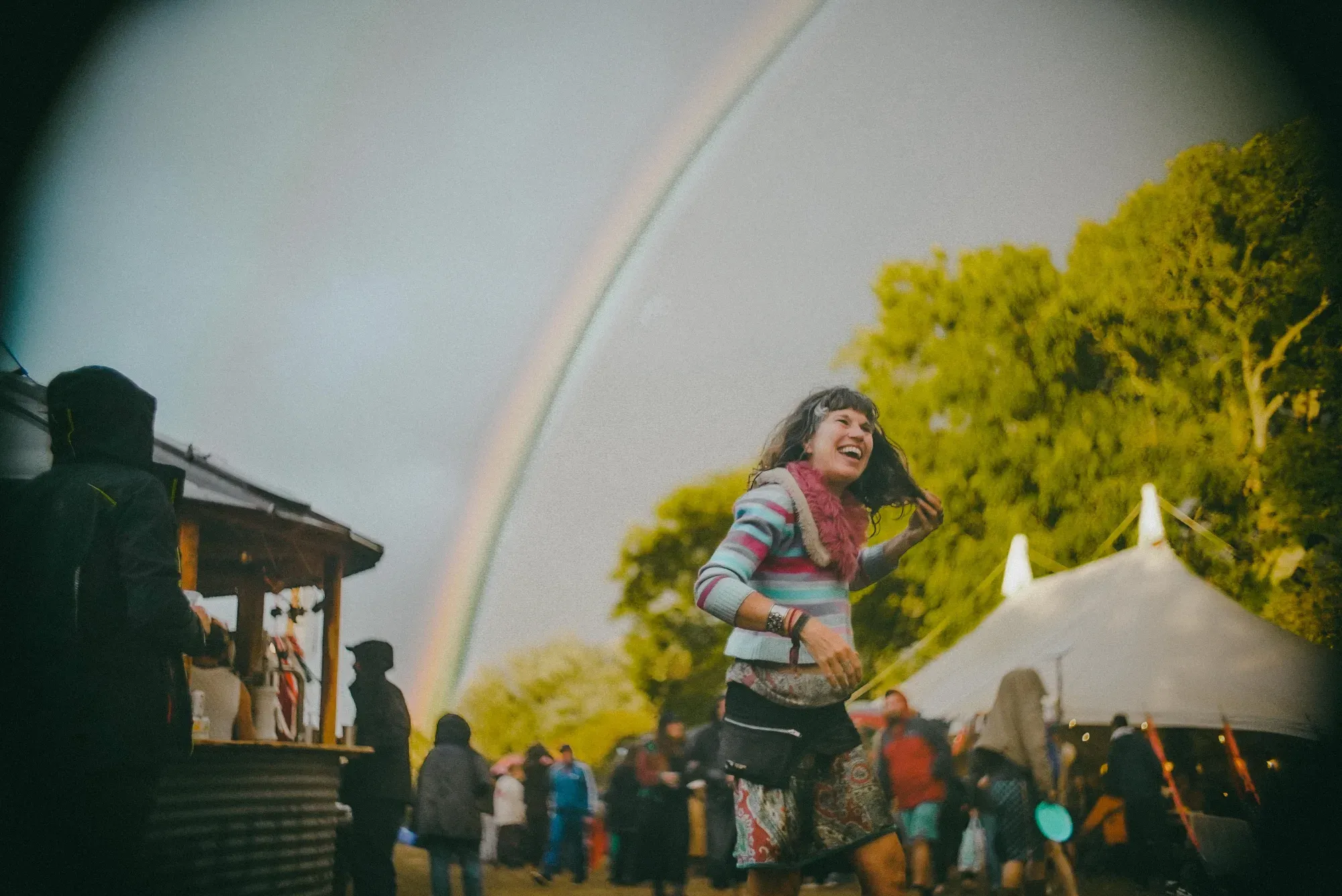 Festival-goer laughing and dancing under a double rainbow, with marquees and food stalls behind.