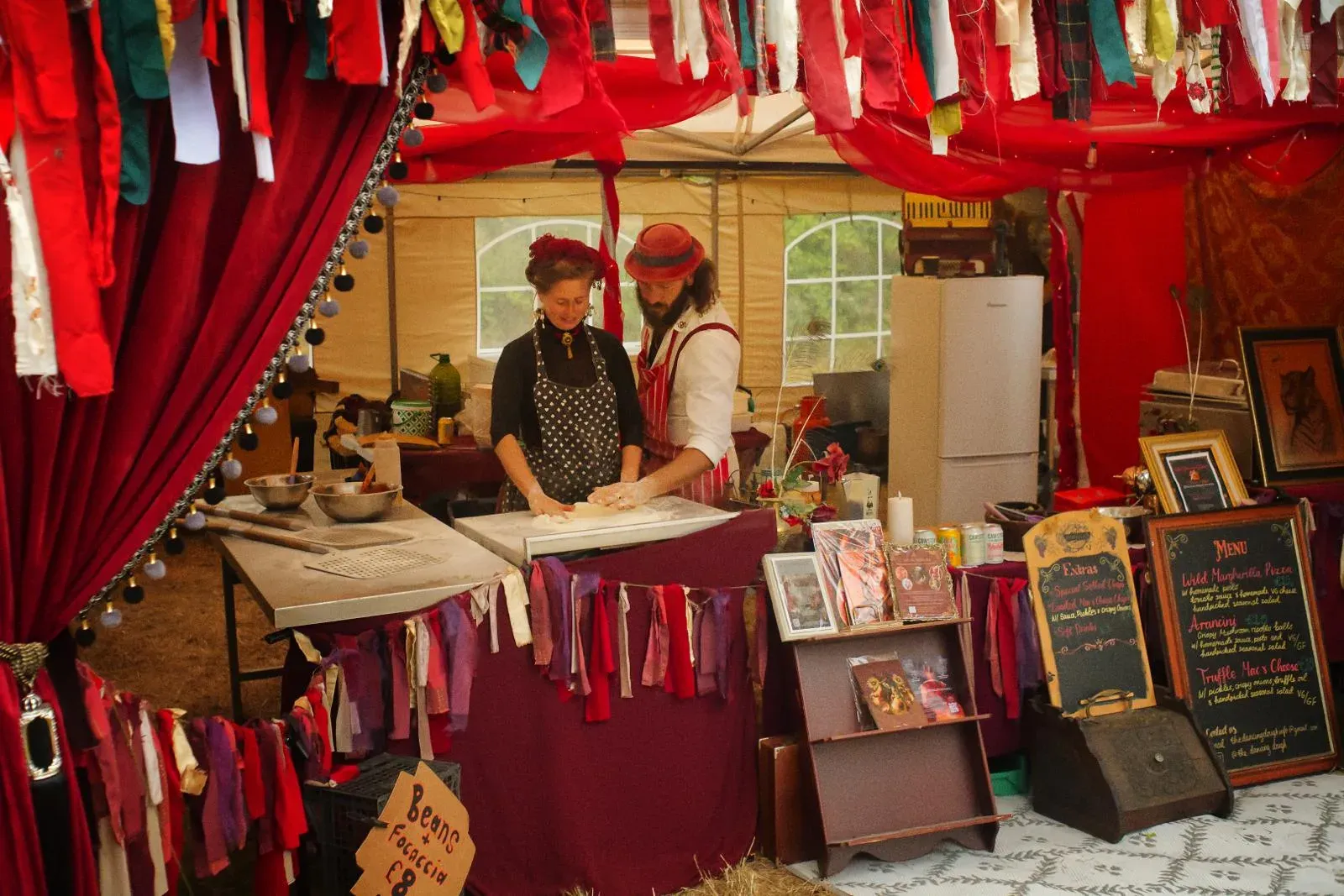 Two costumed vendors hand-making dough at a decorated pizza stall draped in red fabric.