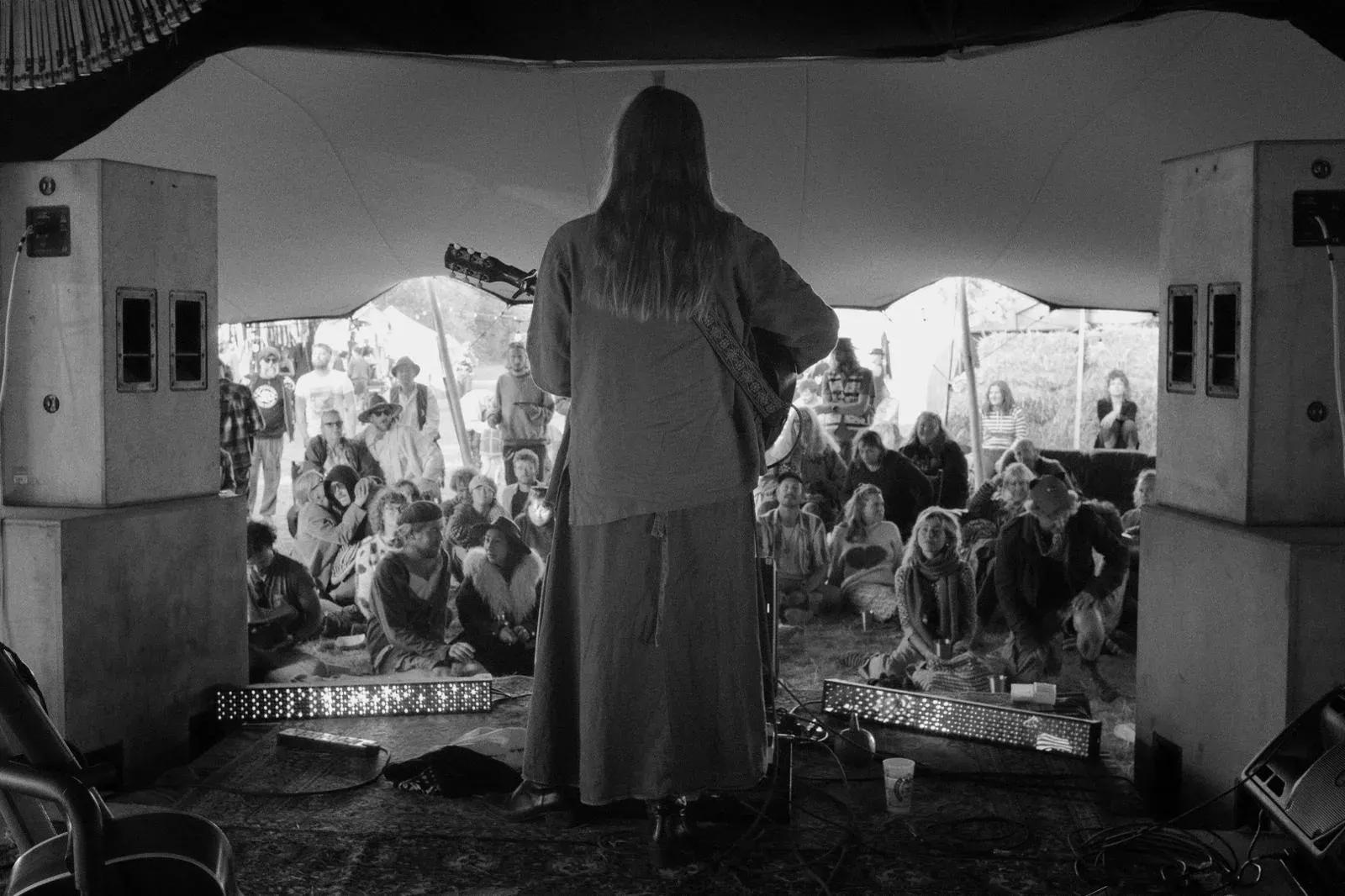Black-and-white view from behind a guitarist on stage, facing a seated, attentive audience under a marquee.