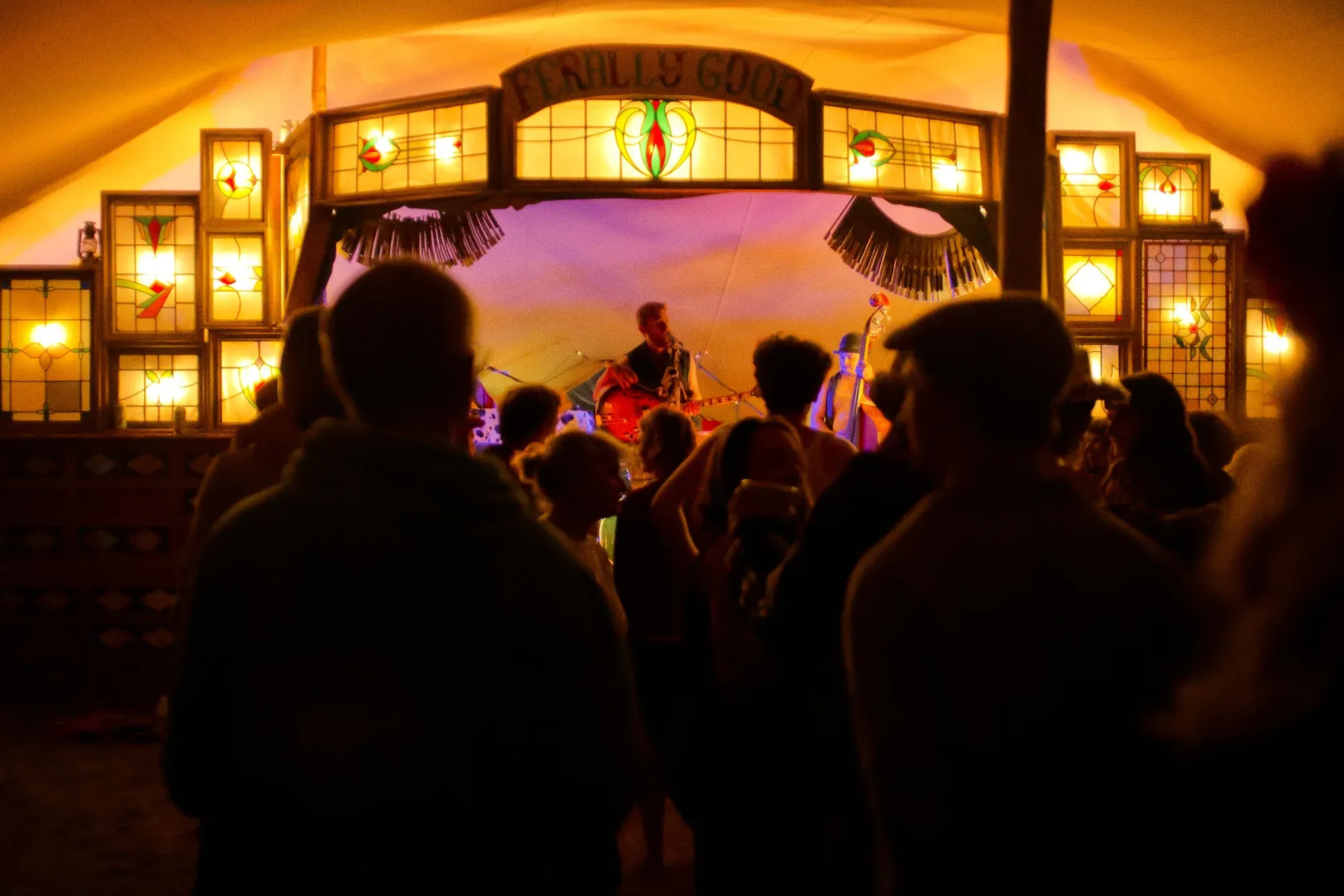 Crowd watching a band on the stained-glass Ferally Good stage, a guitarist and double bassist lit in warm light.