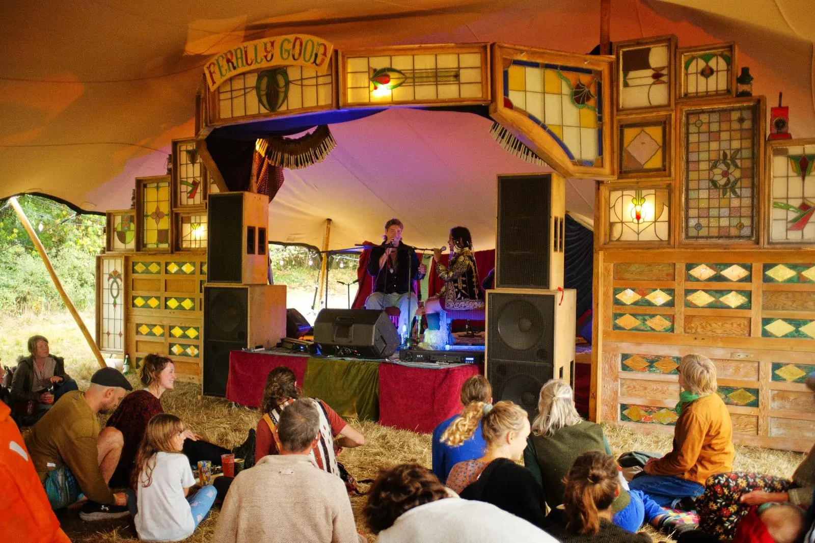 Audience seated on straw watching two performers on the stained-glass Ferally Good stage inside a marquee.