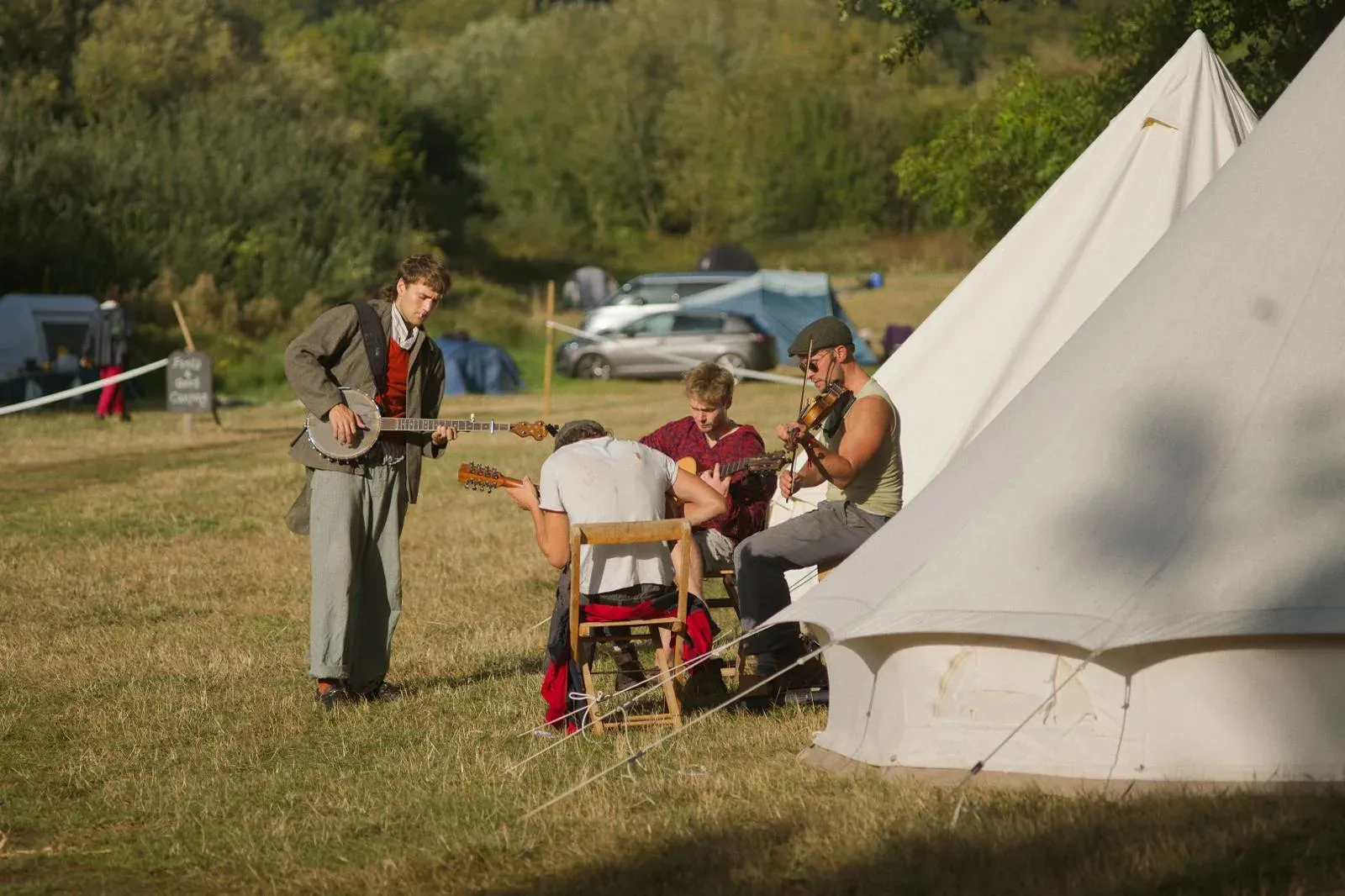 Four musicians playing banjo, mandolin, guitar and fiddle in an impromptu jam by a bell tent at golden hour.