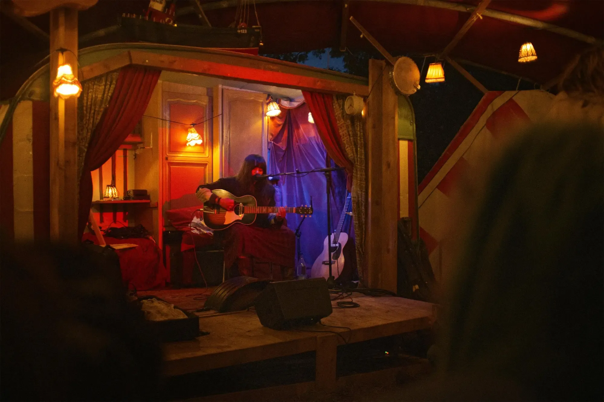 Solo musician playing acoustic guitar on an intimate, lamp-lit vintage stage at night, framed by red curtains.