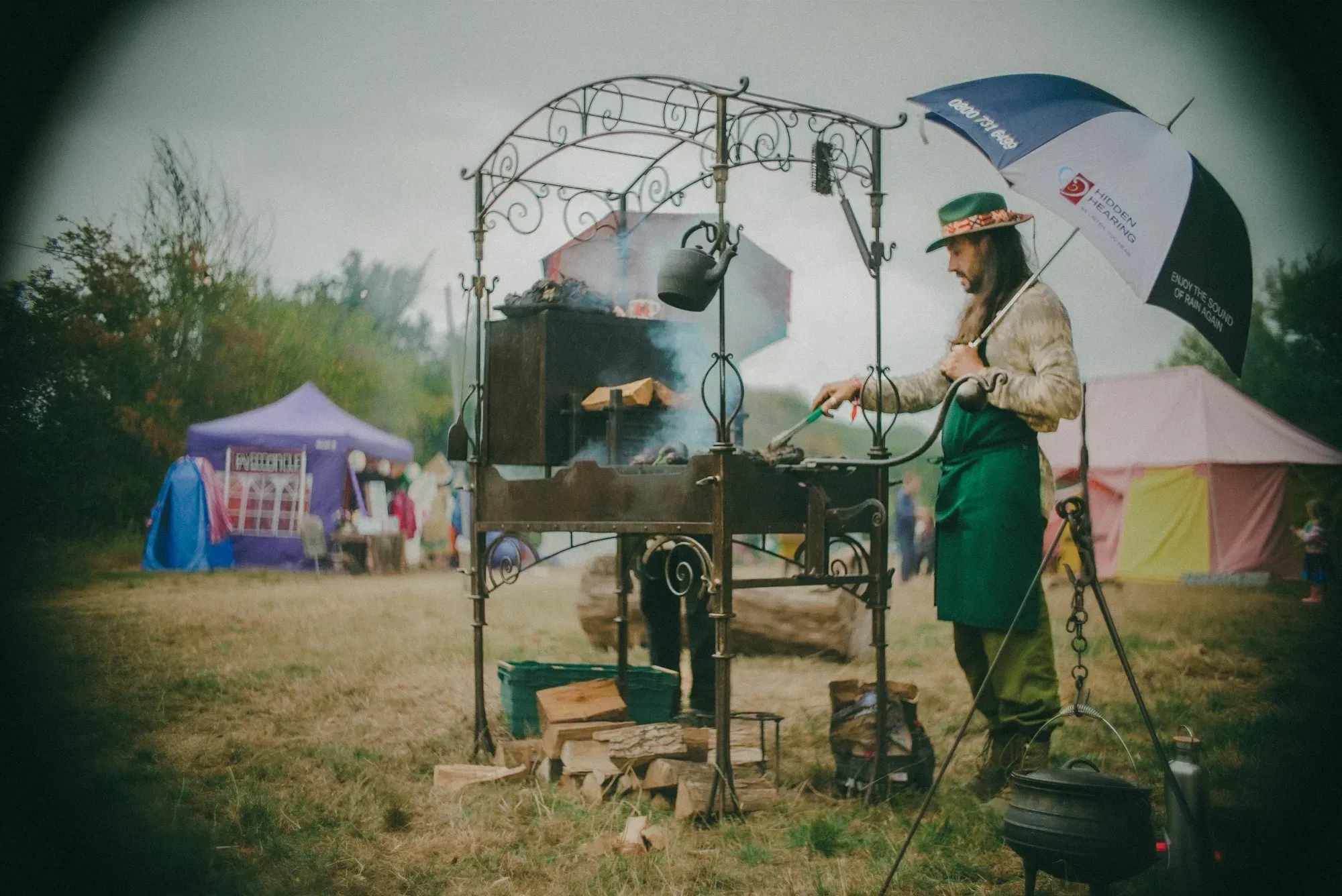 Cook in a green apron tending an ornate wrought-iron wood-fired stove in a field, sheltering under an umbrella.